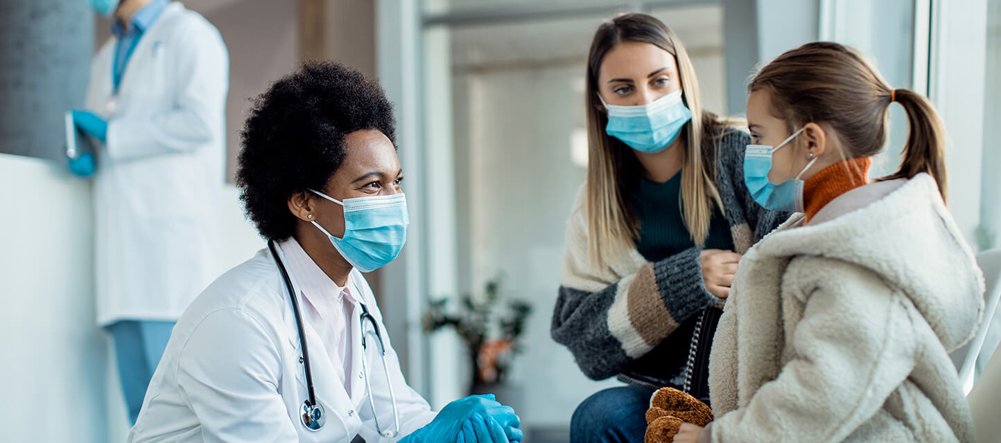 physician talking with mother and daughter in waiting area of hospital while wearing protective masks