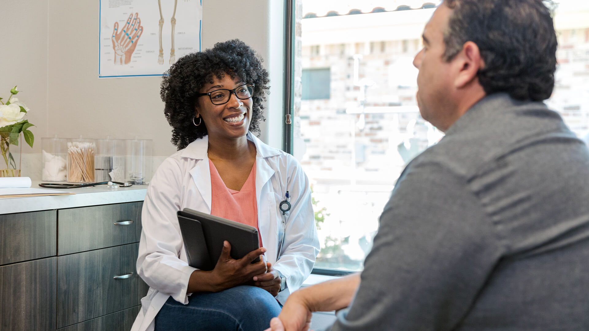 Smiling doctor in a white coat holding a tablet while talking with a seated male patient in an exam room