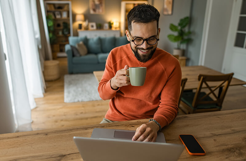 Smiling man with glasses holding a coffee mug while working on a laptop at his dining table at home