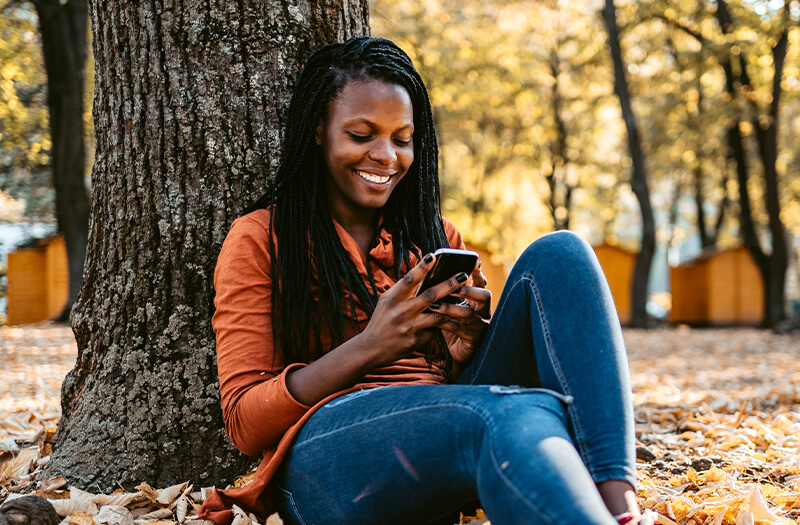 woman of color sitting against a tree checking her phone